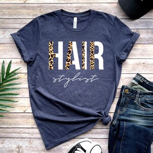 May include: A navy blue t-shirt with the text "HAIR stylist" in white letters. The letters are decorated with a leopard print pattern.