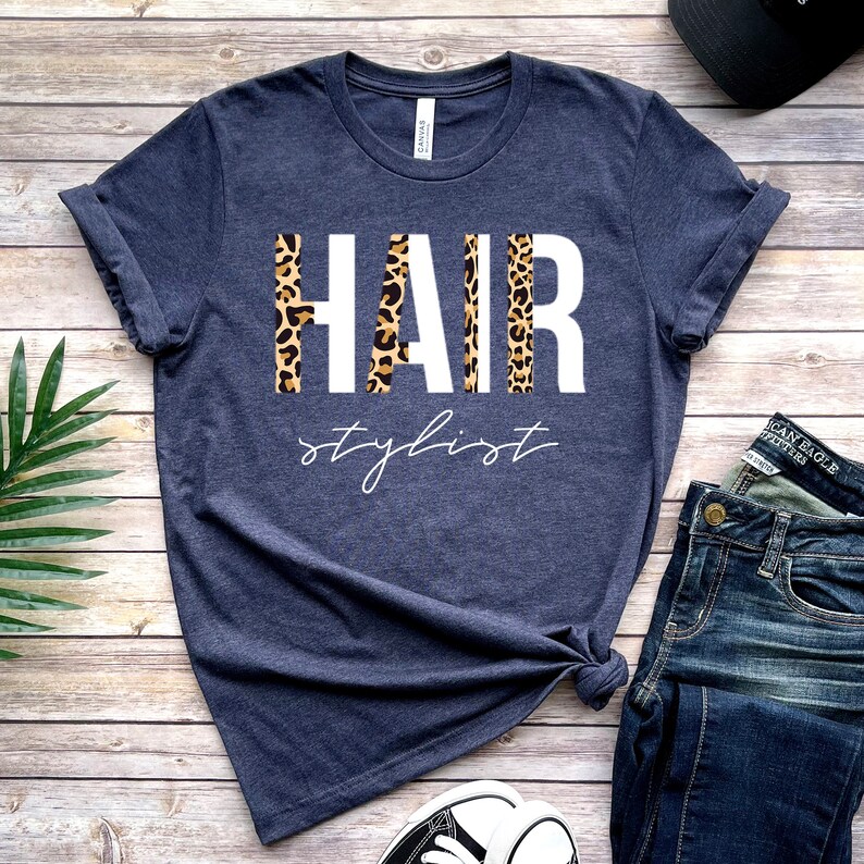 May include: A navy blue t-shirt with the text "HAIR stylist" in white letters. The letters are decorated with a leopard print pattern.