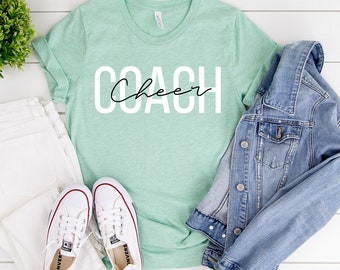 Peut inclure: Un t-shirt vert menthe avec le mot "COACH" en grandes lettres blanches et le mot "Cheer" en écriture cursive. Le t-shirt est associé à un jean blanc, une veste en jean et des baskets blanches.