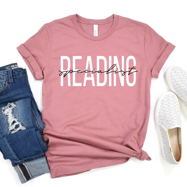 Puede incluir: Una camiseta rosa polvorienta con el texto "READING specialist" en blanco con escritura cursiva negra.