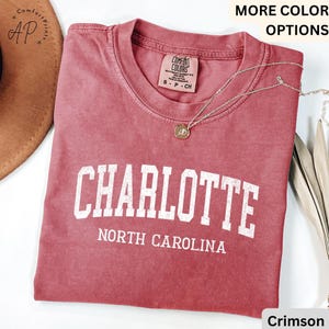 Op de afbeelding: Karmizijnrood T-shirt met het woord "CHARLOTTE" in witte blokletters boven "NORTH CAROLINA". Een zilveren ketting met een hanger is over het shirt gedrapeerd. Het shirt is gemaakt van Comfort Colors materiaal.