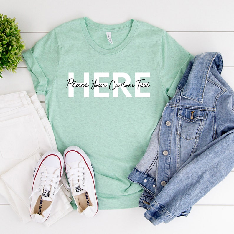 Puede incluir: Una camiseta verde menta con el texto "HERE Place Your Custom Text" en blanco y negro. La camiseta se combina con vaqueros blancos, una chaqueta vaquera y zapatillas de lona blancas con detalles rojos. Se ve una peque&ntilde;a planta verde.