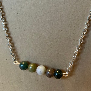 May include: A silver chain necklace featuring five round, multicolored beads. The beads are in shades of green, white, and brown. The necklace has a simple, elegant design.
