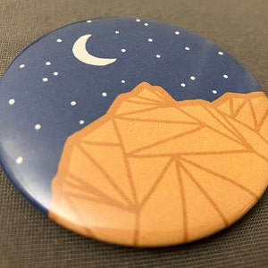 May include: A round button with a blue background and a crescent moon in the upper left corner. The moon is white and has a white outline. The background is dotted with white stars. The bottom half of the button is a tan mountain range with a geometric pattern.