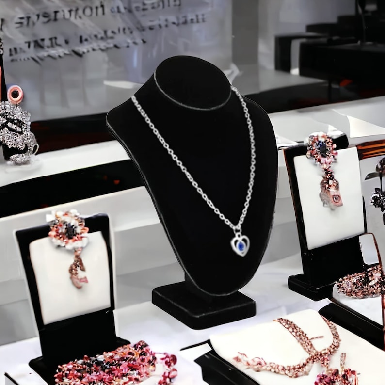 May include: A black velvet necklace display stand with a silver chain necklace with a heart pendant. The necklace is on a white surface with other jewelry displays.