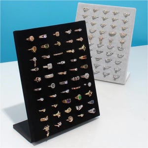 May include: Two ring display stands, one black and one gray, showcasing a variety of rings. The black stand displays gold and silver rings with various gemstone settings. The gray stand features gold and silver rings. The rings are arranged in rows.