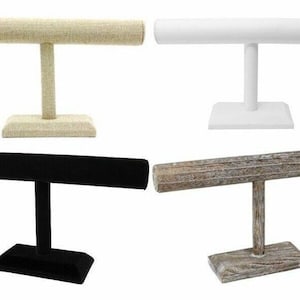 May include: Four jewelry display stands. One is beige linen, one is white, one is black velvet, and the last is a weathered wood finish. Each stand has a horizontal bar supported by a vertical post and a square base.