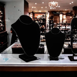 May include: Three black velvet necklace display stands on a white countertop. The stands are different sizes and are all empty.