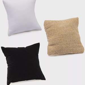 May include: Three small square pillows in different materials. One is white, one is a natural burlap color, and the third is black velvet. The pillows are likely for displaying jewelry or small items.