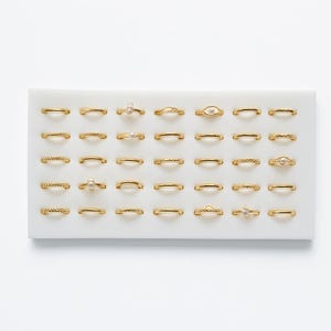 May include: A selection of gold-coloured rings, each with a unique design, including bands with textured patterns and small pearl or gemstone accents. The rings are arranged on a white tray, showcasing a variety of styles and sizes.