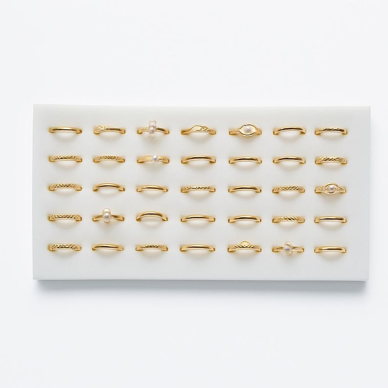 May include: A selection of gold-coloured rings, each with a unique design, including bands with textured patterns and small pearl or gemstone accents. The rings are arranged on a white tray, showcasing a variety of styles and sizes.