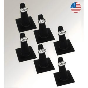 May include: Six black velvet ring displays, each with a silver ring featuring the word "SOLD" in a white oval. The displays are arranged on a white background. An American flag is in the upper right corner.