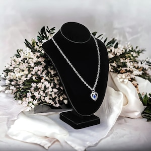 May include: A silver necklace with a heart-shaped pendant featuring a blue gemstone. The necklace is displayed on a black velvet stand.