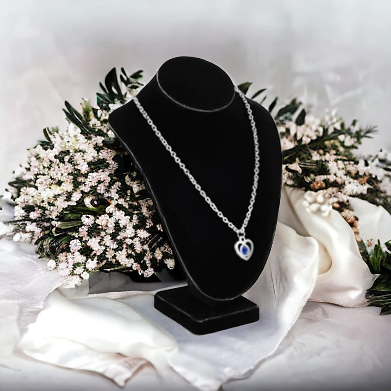 May include: A silver necklace with a heart-shaped pendant featuring a blue gemstone. The necklace is displayed on a black velvet stand.