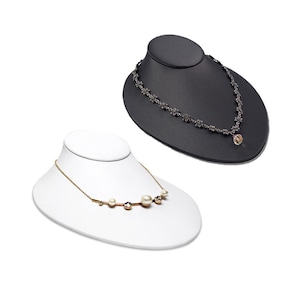 May include: Two necklace display stands, one black and one white, each with a necklace on it. The necklace on the black stand has a silver chain with a small crystal pendant. The necklace on the white stand has a gold chain with a row of pearls and crystals.