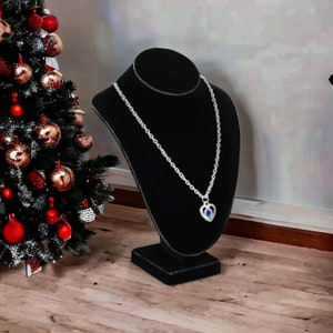 May include: A silver necklace with a heart pendant featuring a blue gemstone on a black velvet jewelry display stand.