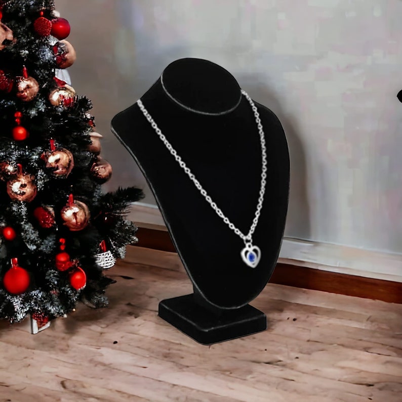 May include: A silver necklace with a heart pendant featuring a blue gemstone on a black velvet jewelry display stand.