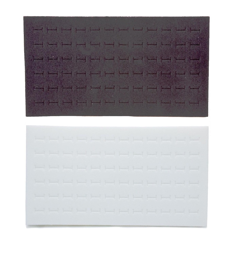 May include: Two rectangular jewellery display inserts. One is dark grey, the other is white. Both have rows of small, evenly spaced slots designed to hold rings or other small jewellery items. The inserts are likely made of foam or a similar material.