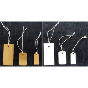 May include: A collection of rectangular and rounded-corner tags in various sizes. Some tags are a light tan color, while others are white. Each tag has a small hole at the top and is attached to a white string. The tags are arranged against a dark textured background.