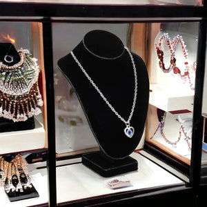 May include: A silver necklace with a blue heart pendant displayed on a black velvet stand in a glass display case. The necklace is on a black velvet stand in a glass display case.