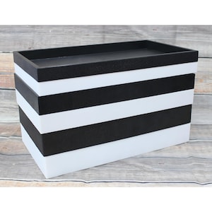 May include: A rectangular storage box with a black top and alternating black and white horizontal stripes. The box has a textured surface and is designed for organizing items. The box is made of a sturdy material.