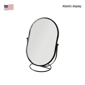 May include: Oval-shaped vanity mirror with a black metal frame and stand. The mirror has a clean, reflective surface. The base is a curved, minimalist design. The text "Atlantic display" is visible.