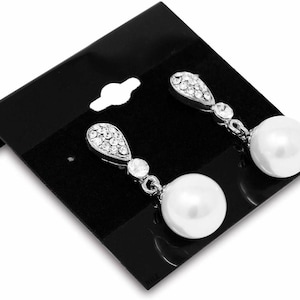 May include: A pair of silver-toned drop earrings with a pearlescent white, round pearl at the bottom. The top of the earrings feature a teardrop-shaped design encrusted with small, clear crystals. The earrings are displayed on a black card.