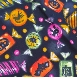 May include: A black fabric featuring a repeating pattern of colorful Halloween-themed candies. The print includes orange jack-o'-lanterns, green spiders, pink witches on broomsticks, orange bats, and green robots, interspersed with small yellow and pink dots.