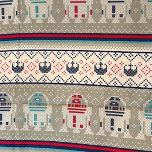 Könnte beinhalten: Ein Stoff mit einem sich wiederholenden, verpixelten Muster aus Droiden in Rot, Blau und Hellblau auf beigem Hintergrund. Das Design umfasst horizontale Streifen mit einem geometrischen Rautenmuster und dunkelblauen Sternsymbolen auf cremefarbenem Grund.