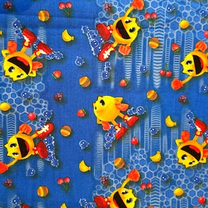 Pode incluir: Um tecido azul com um padrão repetitivo de personagens Pac-Man amarelos usando tênis vermelhos, intercalados com pequenas ilustrações de cerejas, bananas, morangos, uvas e itens de jogo redondos coloridos sobre um design de colmeia.