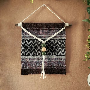 May include: A woven wall hanging with a brown, black, and white geometric pattern. A white cord with three wooden beads, two green and one natural, hangs from the center of the hanging.