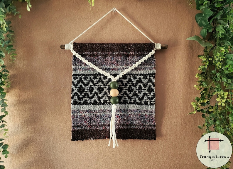 May include: A woven wall hanging with a brown, black, and white geometric pattern. A white cord with three wooden beads, two green and one natural, hangs from the center of the hanging.