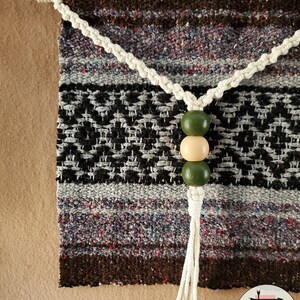 May include: A woven wall hanging with a brown, gray, and black pattern. The hanging has a white cord with a tassel and three wooden beads in green and beige.