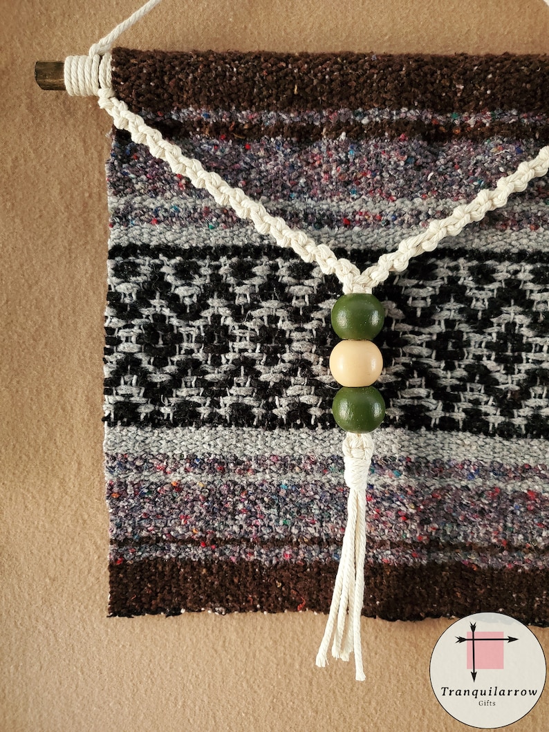 May include: A woven wall hanging with a brown, gray, and black pattern. The hanging has a white cord with a tassel and three wooden beads in green and beige.