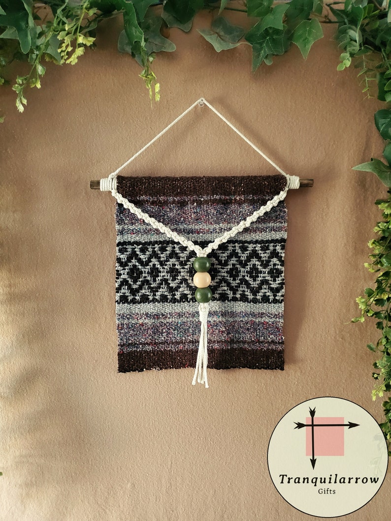 May include: A woven wall hanging with a brown, black, and white geometric pattern. The hanging has a white tassel and a string of wooden and green beads.