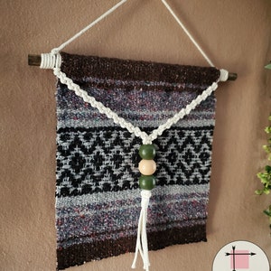 May include: A brown, black, and white woven wall hanging with a white tassel and three wooden beads. The beads are green and natural wood.