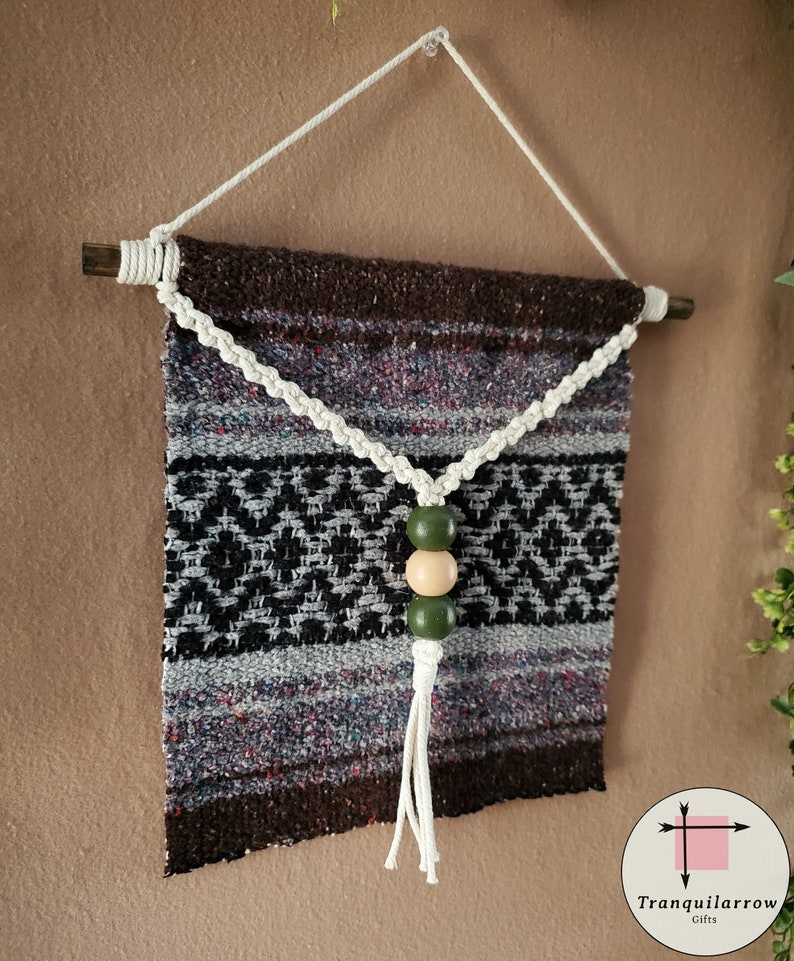 May include: A brown, black, and white woven wall hanging with a white tassel and three wooden beads. The beads are green and natural wood.