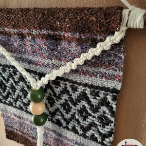 May include: A brown, white, and black woven wall hanging with a white rope and green and beige beads. The hanging is attached to a wooden dowel.