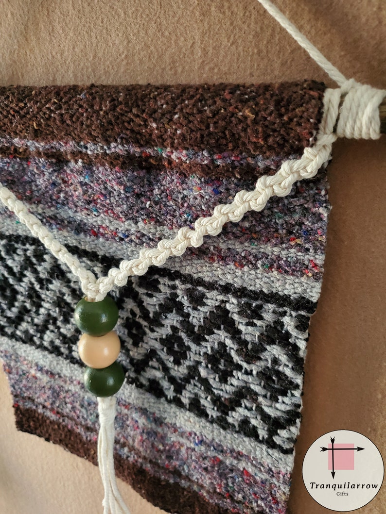 May include: A brown, white, and black woven wall hanging with a white rope and green and beige beads. The hanging is attached to a wooden dowel.
