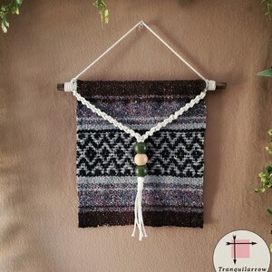 May include: A brown and gray woven wall hanging with a white macrame tassel and three wooden beads. The beads are green and beige. The hanging is attached to a wooden dowel rod.