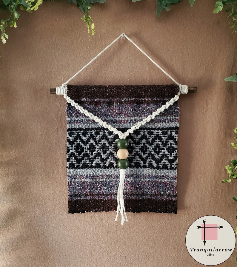 May include: A brown and gray woven wall hanging with a white macrame tassel and three wooden beads. The beads are green and beige. The hanging is attached to a wooden dowel rod.