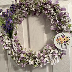 May include: A purple and white fabric wreath with a small bouquet of purple flowers on the left side. A tag with a watercolor painting of a bouquet of yellow flowers in a glass vase is attached to the wreath.