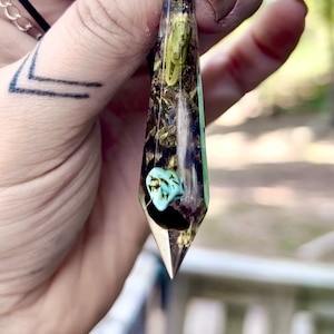 May include: A clear, pointed pendant with a silver bail. The pendant is filled with dried herbs and flowers, and a small turquoise stone is visible in the center.