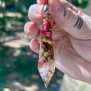 May include: A clear resin pendant with a silver-colored bail. The pendant is filled with dried flowers in shades of red, pink, green, and gold. The pendant is held up against a blurred green background.