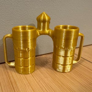 Puede incluir: Un doble mug dorado, impreso en 3D, con un arco de conexión y una parte superior puntiaguda. Cada taza tiene un asa y un cuerpo cilíndrico con crestas horizontales. El diseño es simétrico y parece un artículo decorativo.