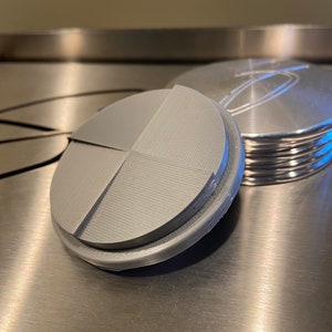May include: Two grey, circular lids. One has a segmented design, the other has ridges and an etched design. Both are metal and rest on a brushed metal surface. The lids are likely for a container.