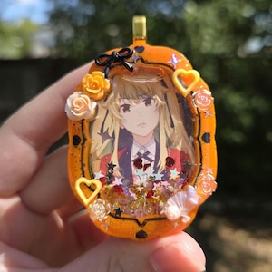 May include: Orange resin pendant with a portrait of an anime character. The pendant is decorated with small flowers, hearts, and stars. A black bow is at the top, and a gold-colored bail is attached for hanging.