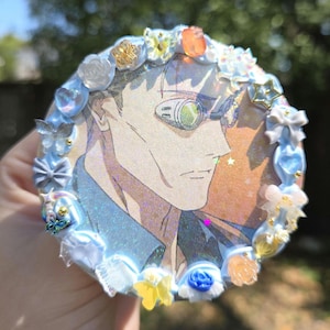 Puede incluir: Un pin redondo y decorativo con un personaje de anime con gafas de color verde. El pin está bordeado con purpurina azul claro y adornado con pequeños lazos, flores y adornos en forma de estrella. El fondo tiene un degradado naranja y azul.