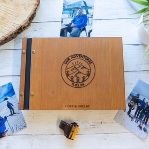 Könnte beinhalten: Ein Holzfotoalbum mit schwarzem Rücken und Messingakzenten. Der Einband ist mit dem Text "Our Adventures 11.05.24" und "Luke & Shelby" graviert.