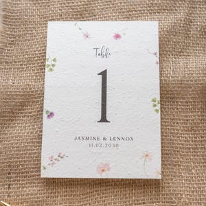 May include: A white table number card featuring the number 1 in a bold, dark font. The card is adorned with small pink and purple floral designs and the text "Table" and "Jasmine & Lennox 11.02.2030". It rests on a burlap surface.